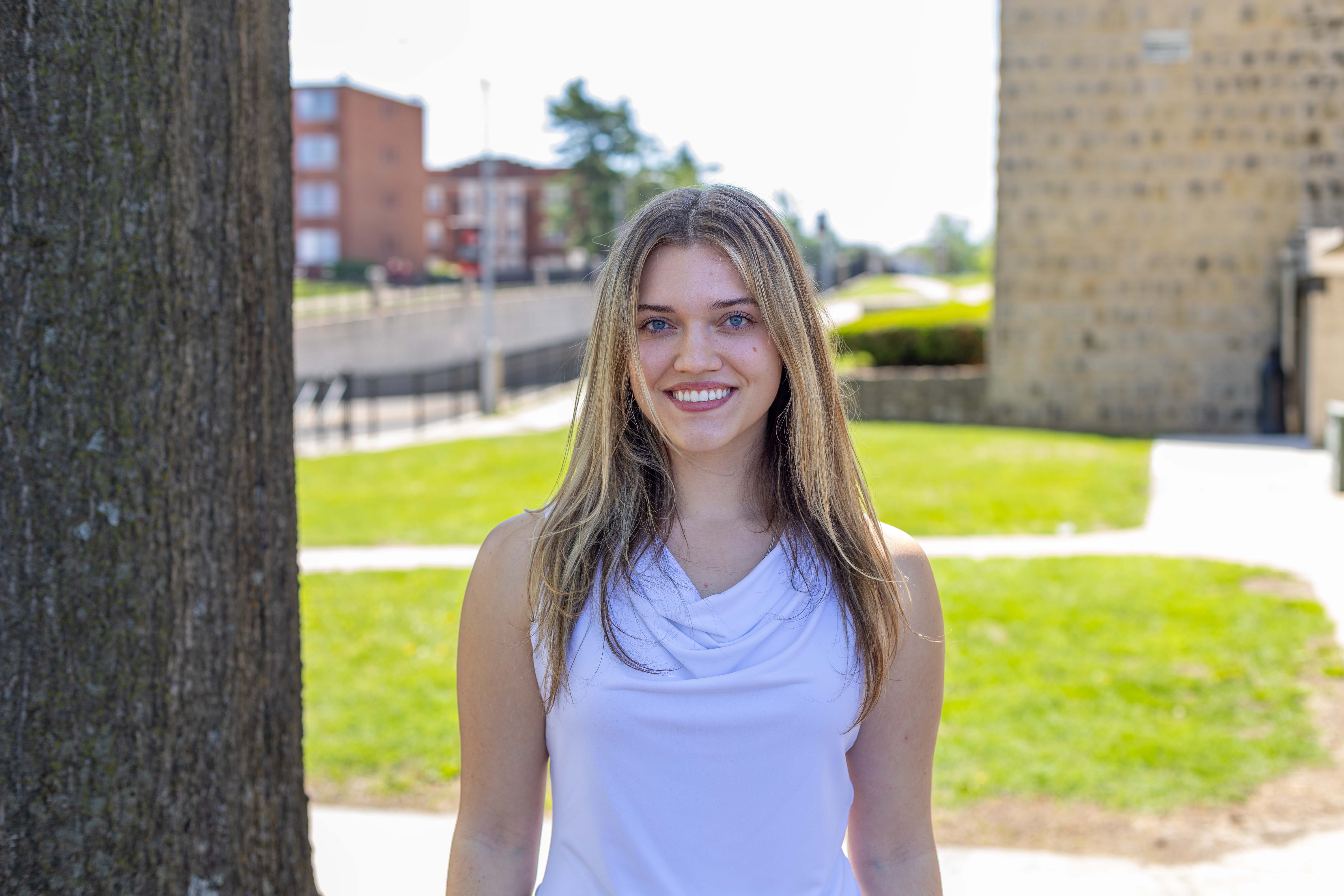 A person is wearing a sleeveless white shirt while standing outside on the UCM campus in a grassy area next to a tree. Campus buildings can be seen in the background.