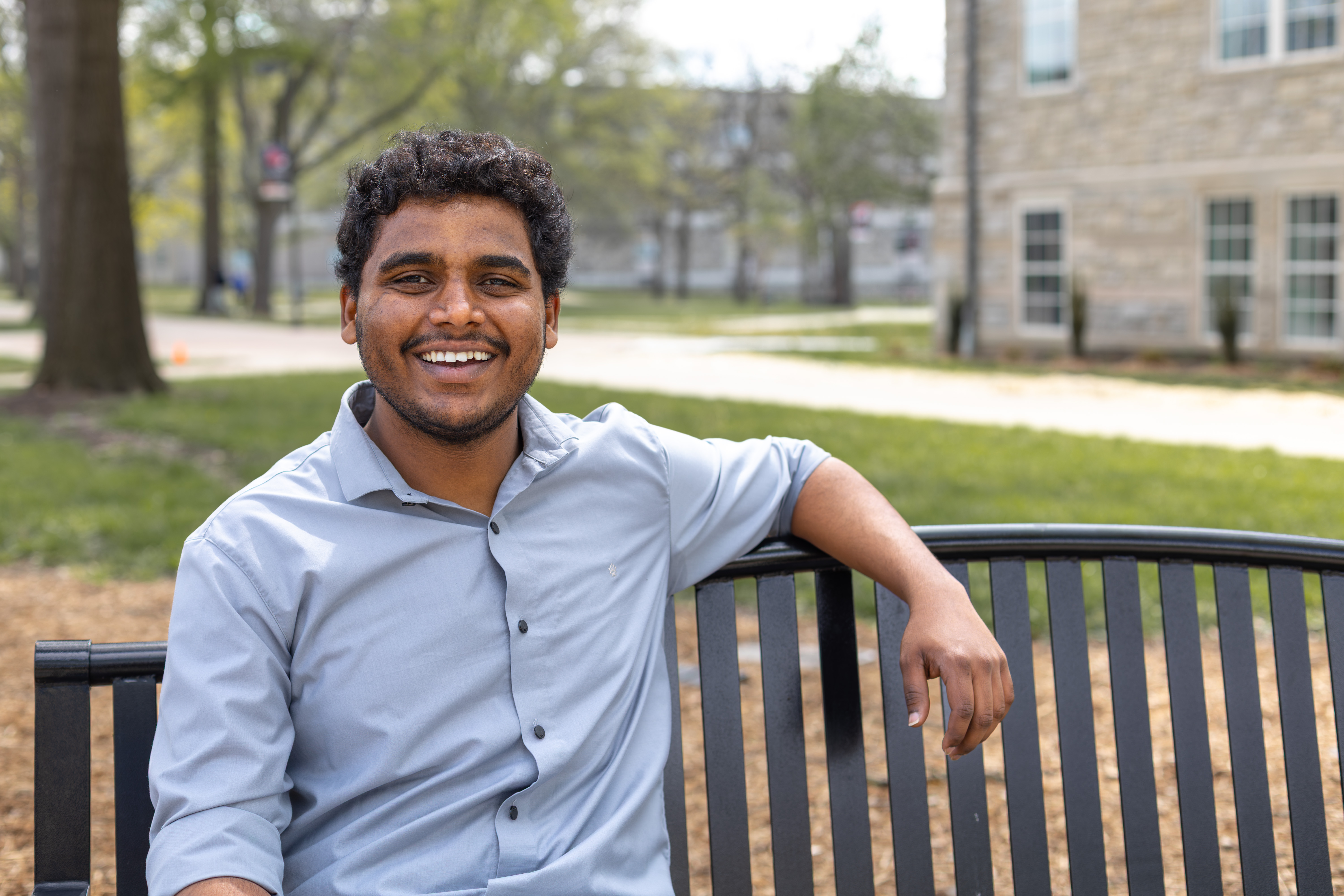A person is wearing a collared light-blue button-up shirt with the sleeves slightly rolled up. They are looking at the camera and smiling while sitting on a bench outside on the UCM campus. Grass, buildings and trees can be seen in the background.