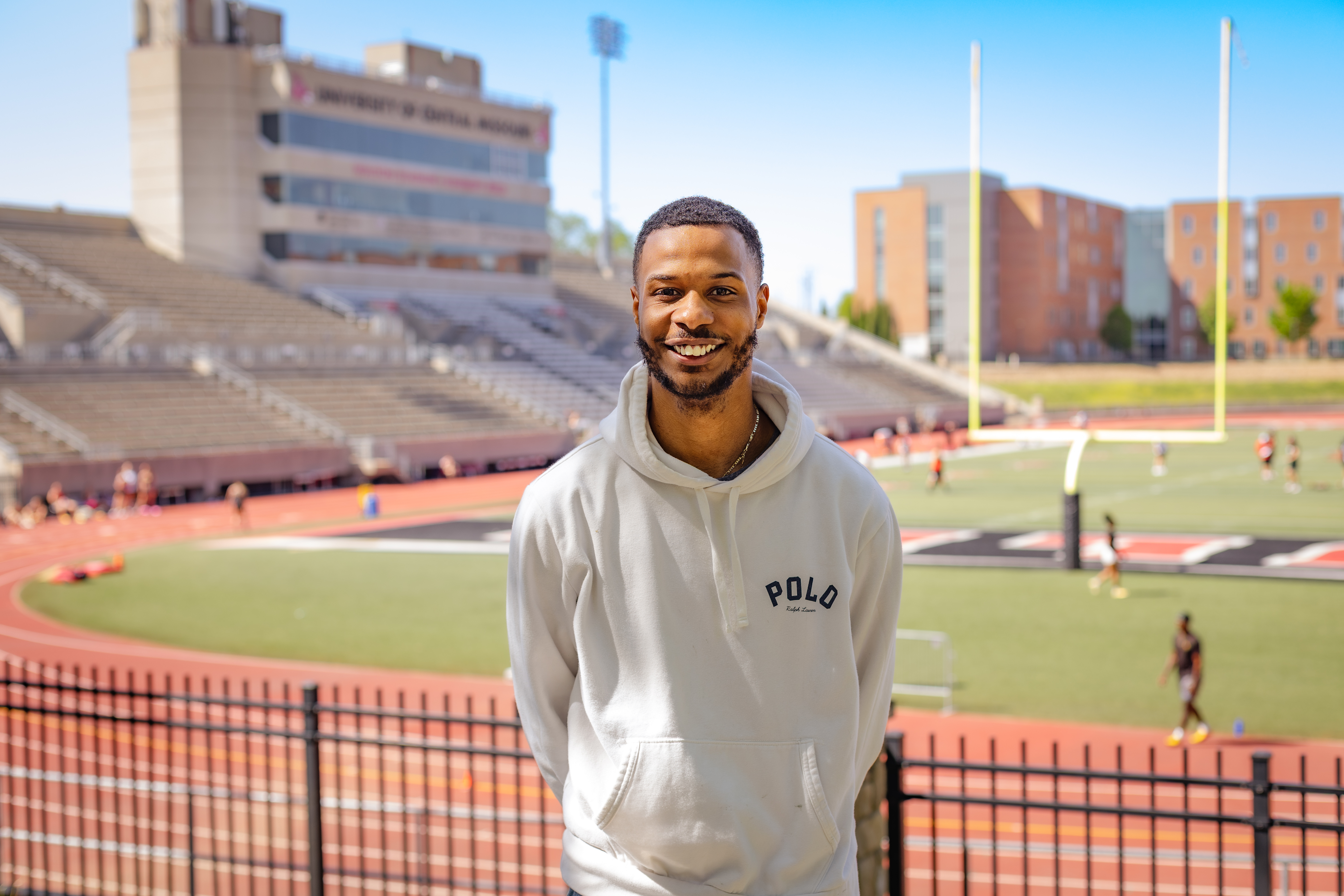 A person is wearing a white hooded sweatshirt. It is sunny outside and behind the person is a football stadium, where people are practicing for various sports.