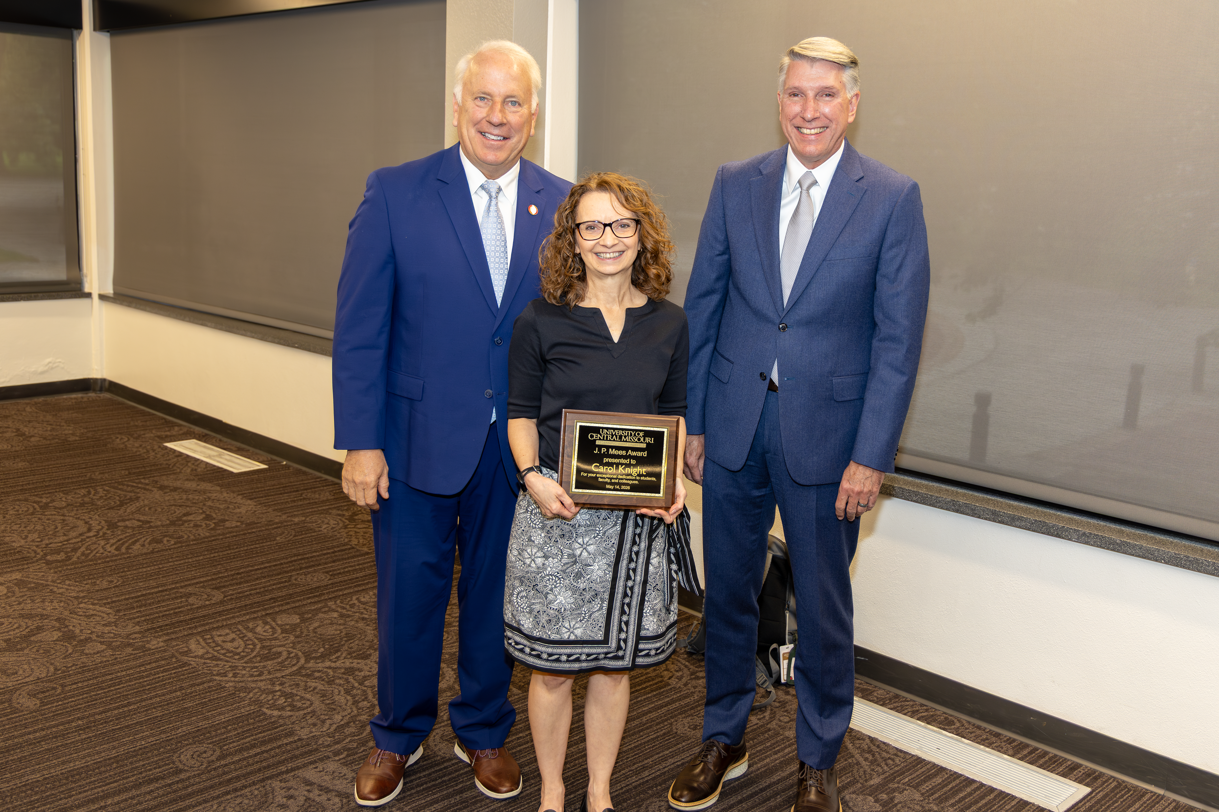 Three people are in a meeting room in the Elliott Student Union. One is in the front and holding an award plaque. The other two are standing behind the award winner.