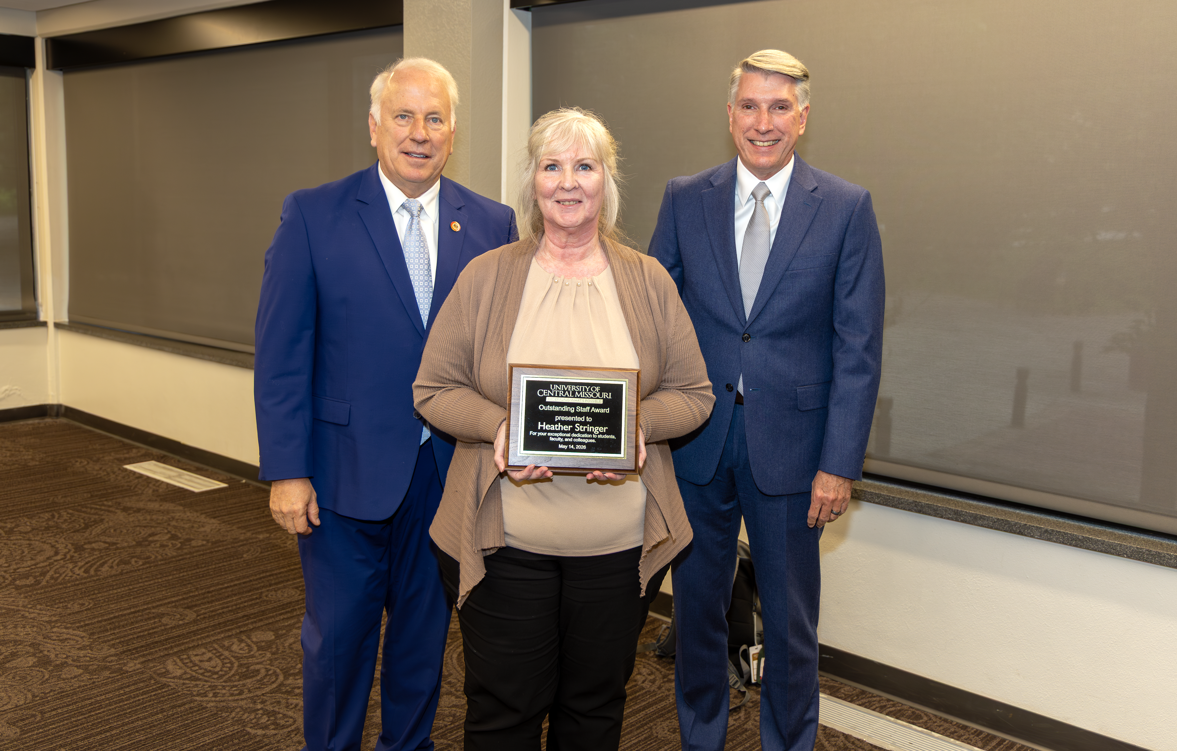 Three people are in a meeting room in the Elliott Student Union. One is in the front and holding an award plaque. The other two are standing behind the award winner.