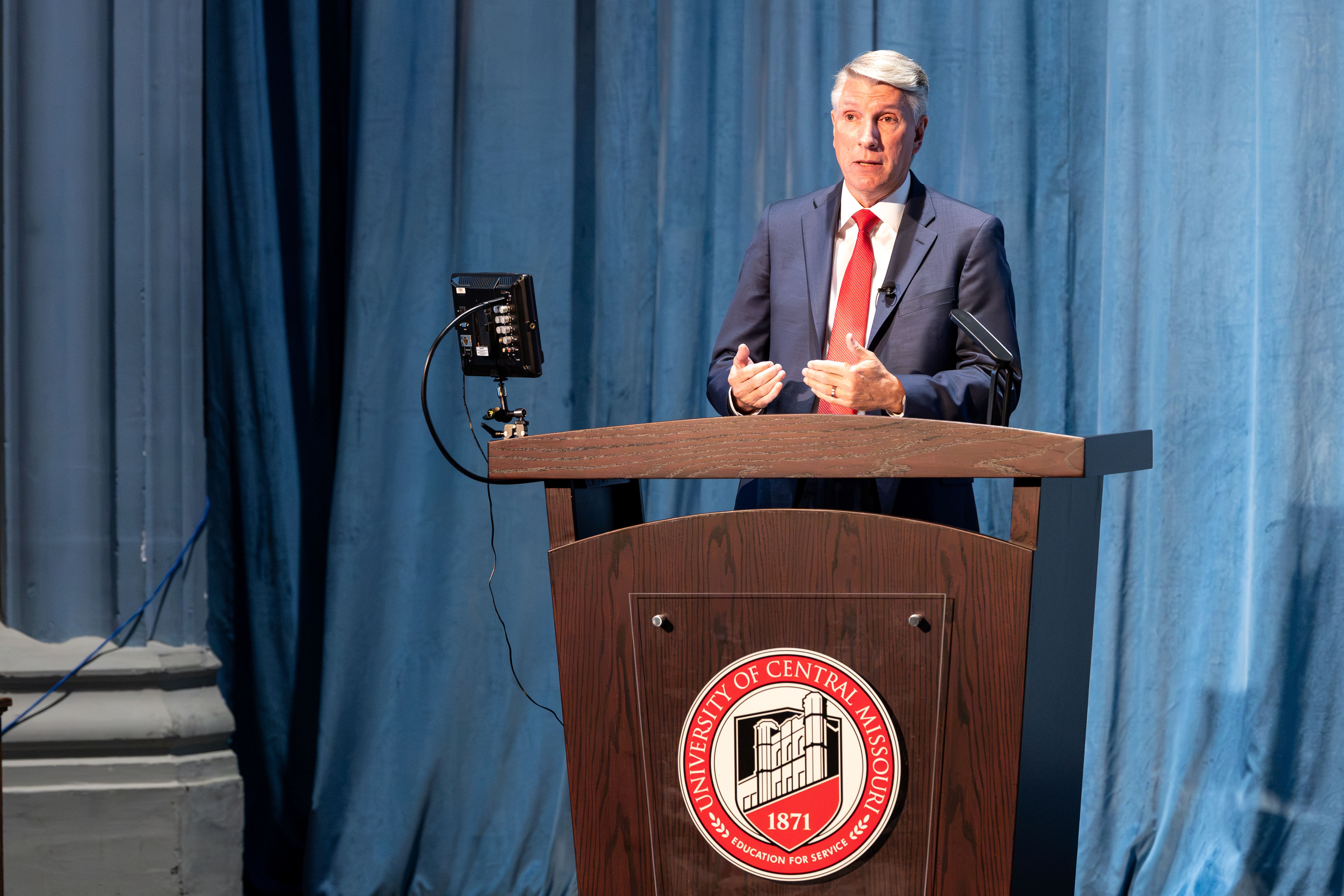 President Roger Best stands behind a brown wooden podium with the UCM logo on the front. He is in front of blue curtains while standing on the Hendricks Hall stage. He is wearing a navy suit, white shirt and red tie. 
