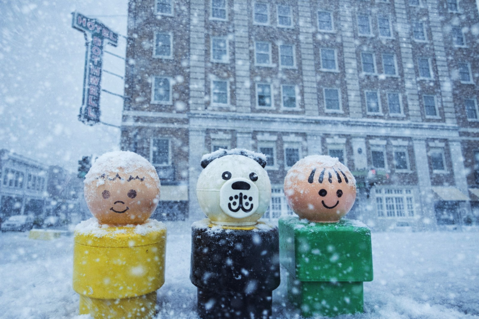 A photo of three children's toys with light snow falling on them. They are "standing" on the sidewalk across from historic Hotel Bothwell in downtown Sedalia, Missouri, while snow falls all around.