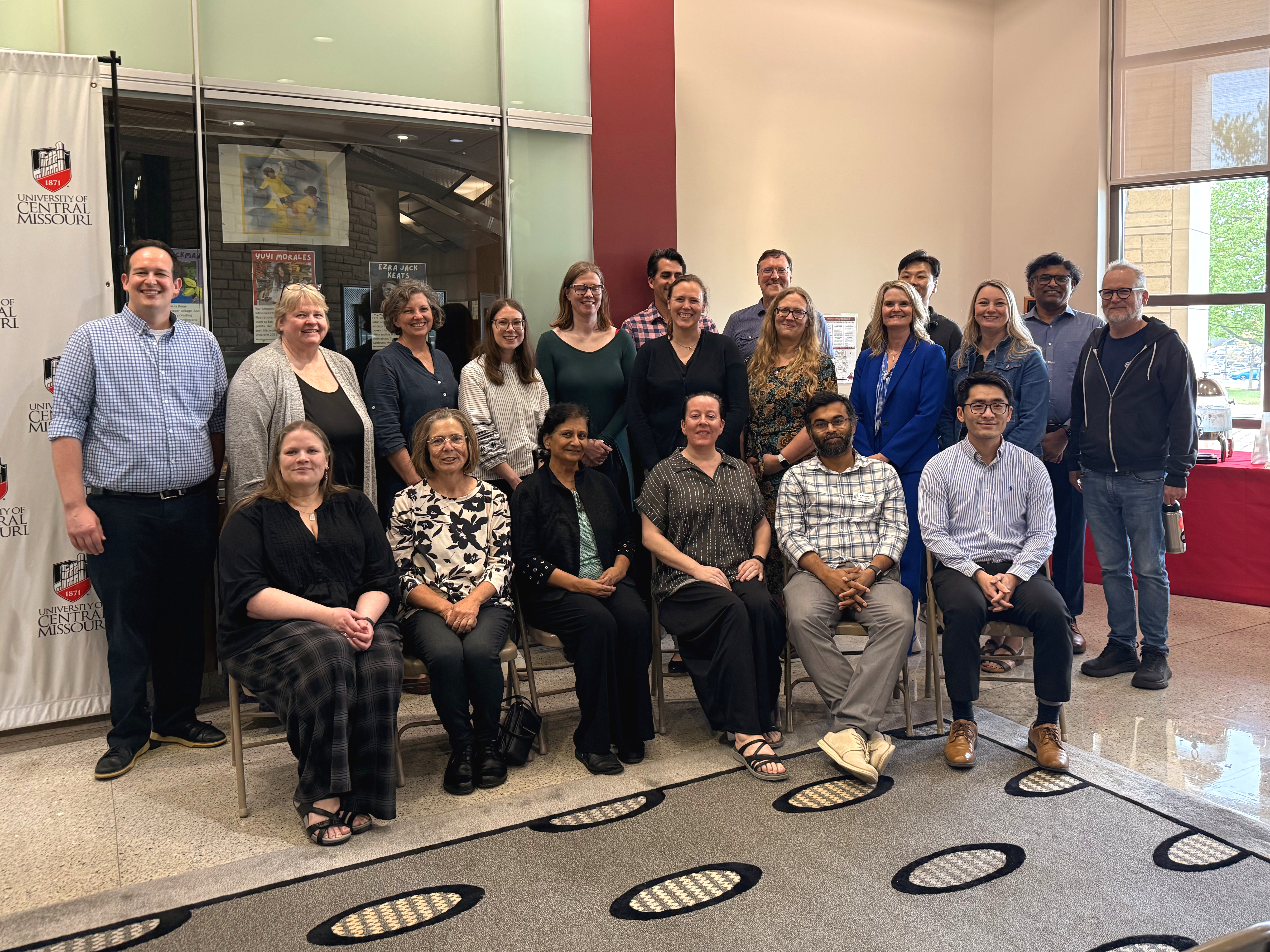 Twenty faculty members pose for a photo on the first floor of the James C. Kirkpatrick Library. Six people are sitting in chairs in the front row, with 10 standing in the second row and four standing in the back row.