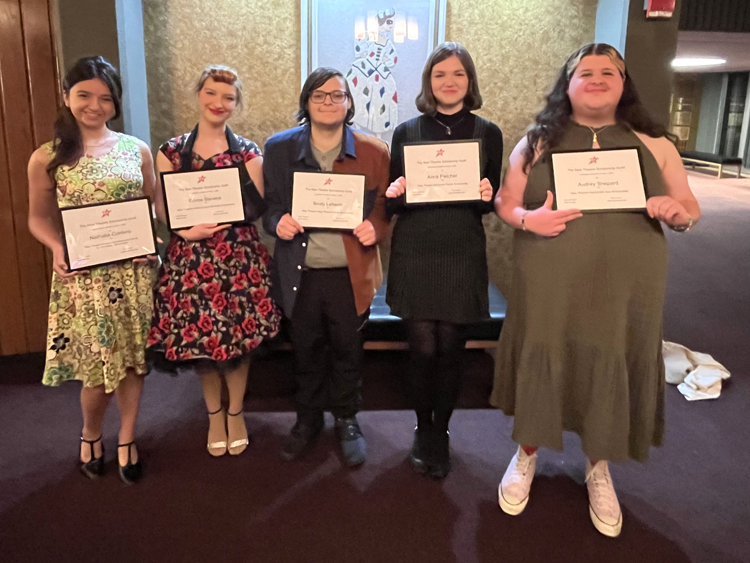 Five students are standing next to each other in the Highlander Theatre lobby. Each is holding a white paper certificate for their New Theatre Guild Scholarship.