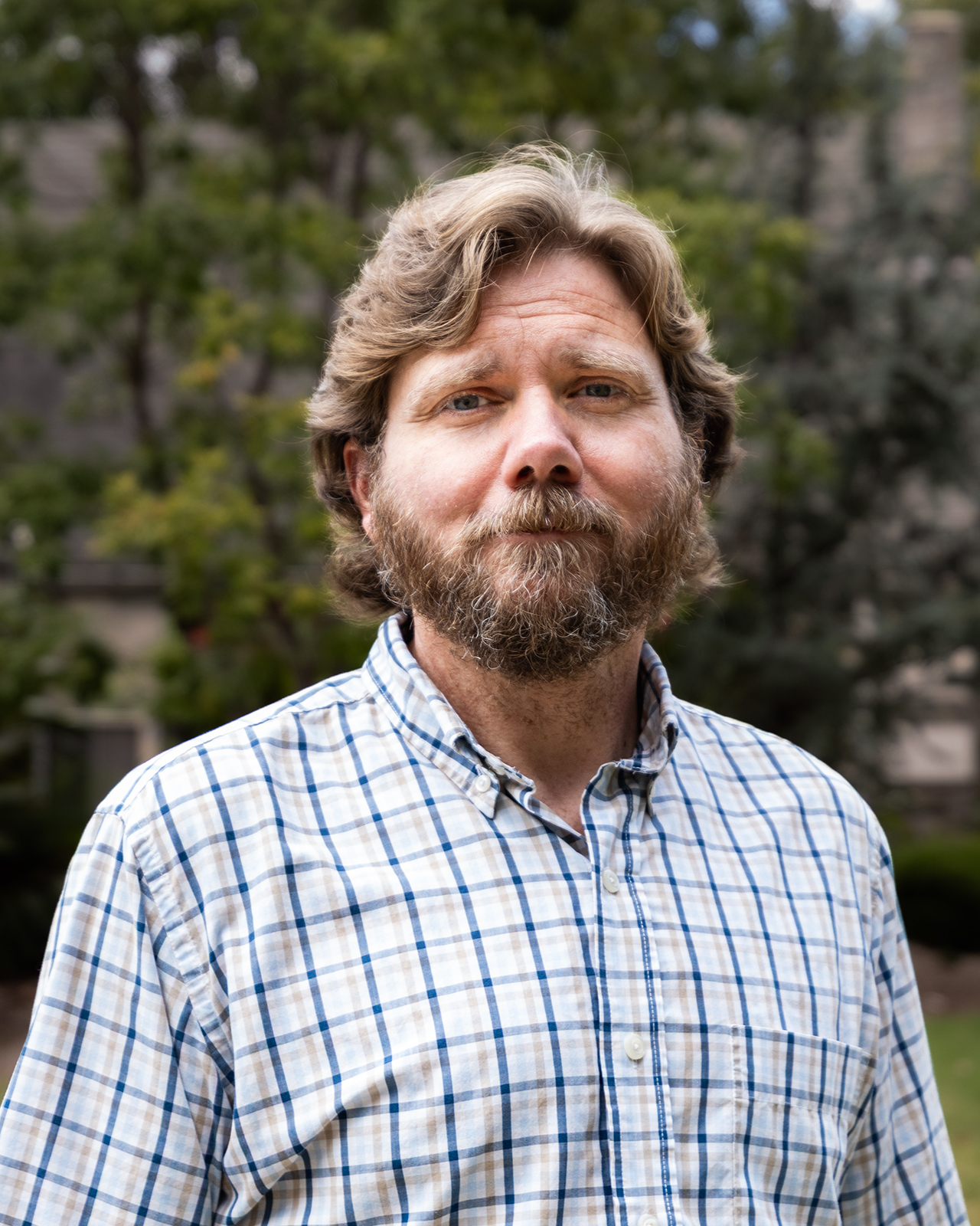 A person with brown hair and a beard is seen in a portrait photo from the shoulders up, wearing a white-and-blue plaid button-up shirt and standing in front of a tree.