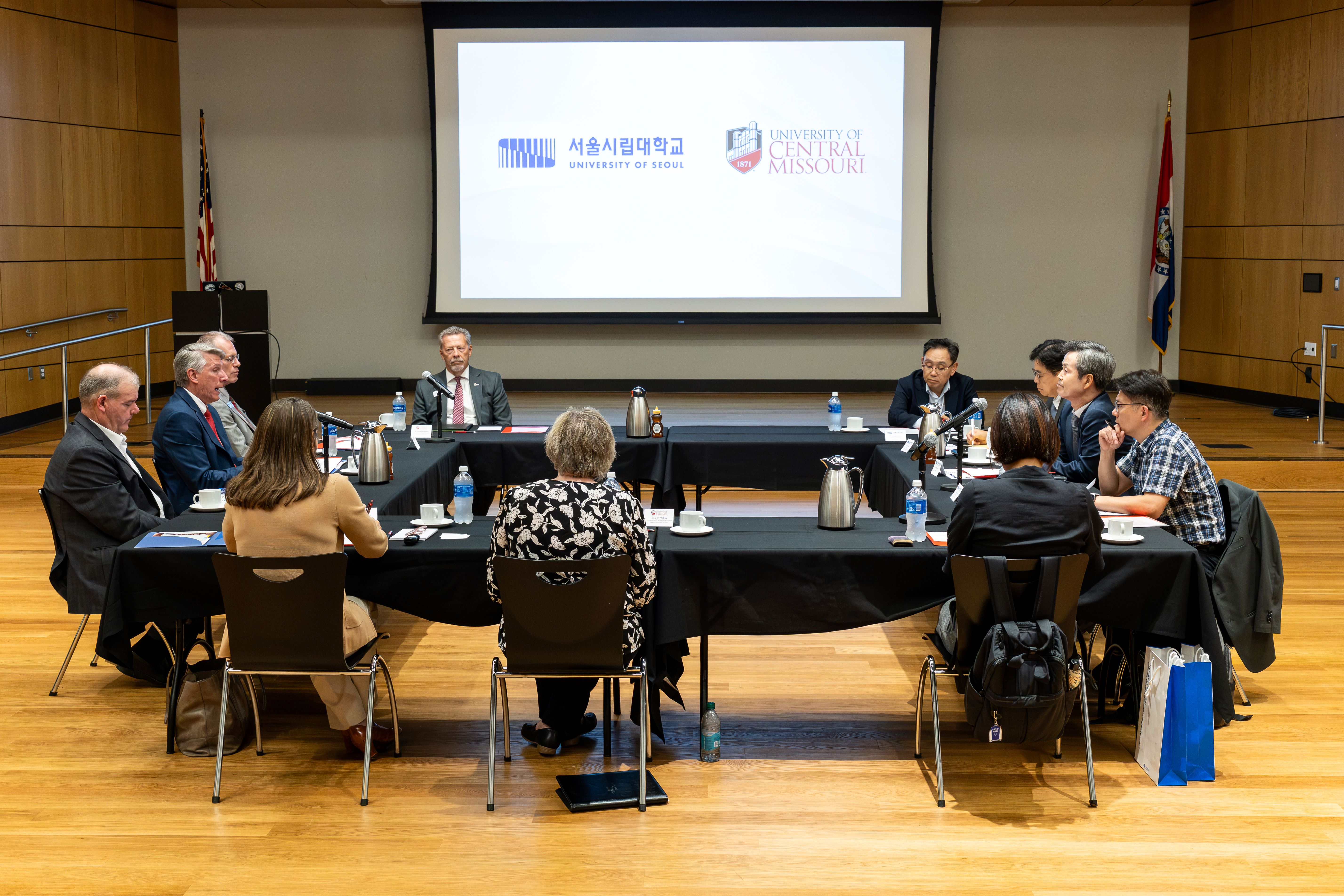 Eleven people sit around long tables that have been arranged into a square and covered with black tablecloths. The projection screen behind them shows the UOS and UCM logos. Ten of the people are listening while one person is talking to the group.