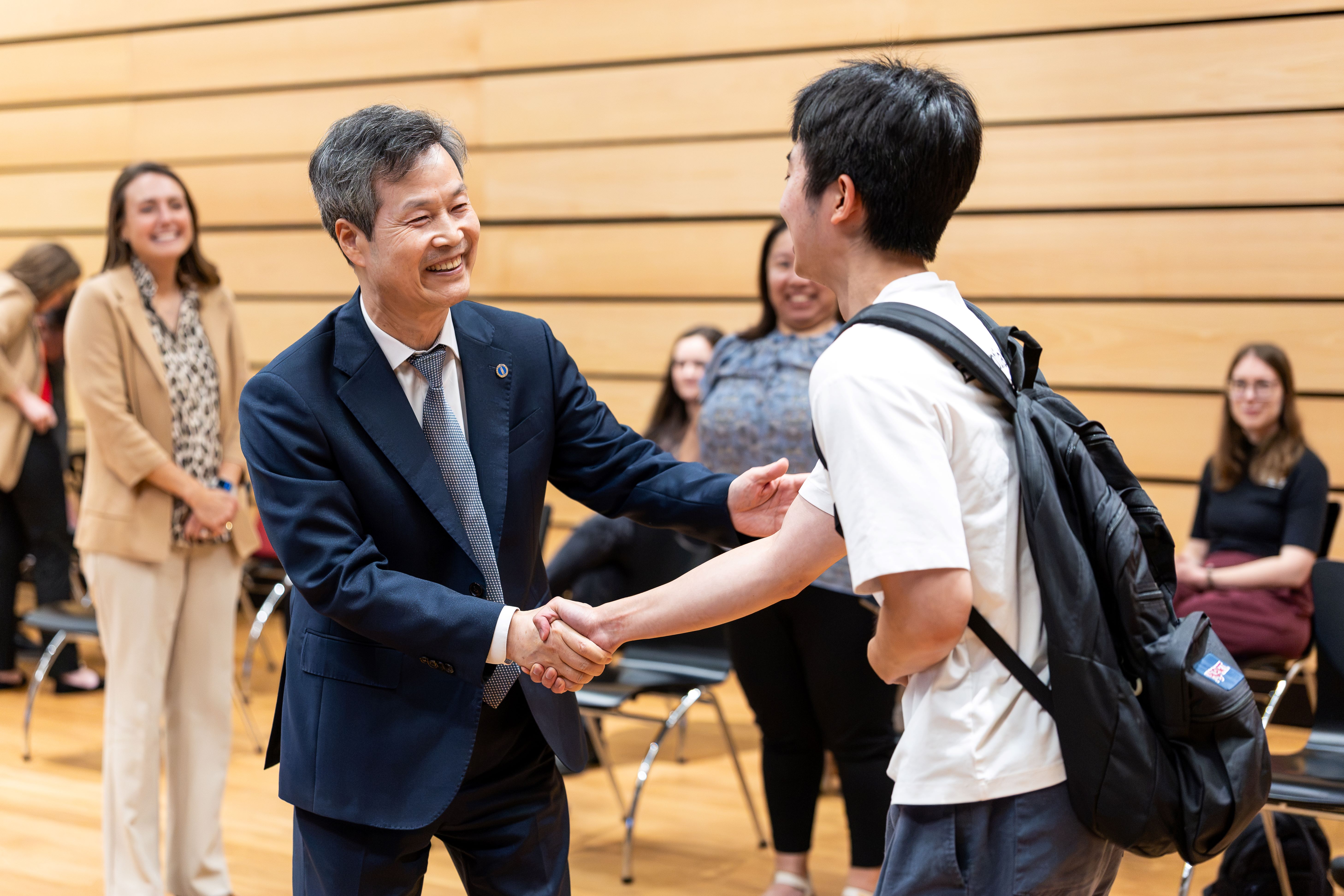 The UOS president shakes hands with a UOS student who is studying at UCM. The president is wearing a navy suit and the student is wearing a white T-shirt and gray/blue pants. People are seen in the background watching the interaction or talking to each other.