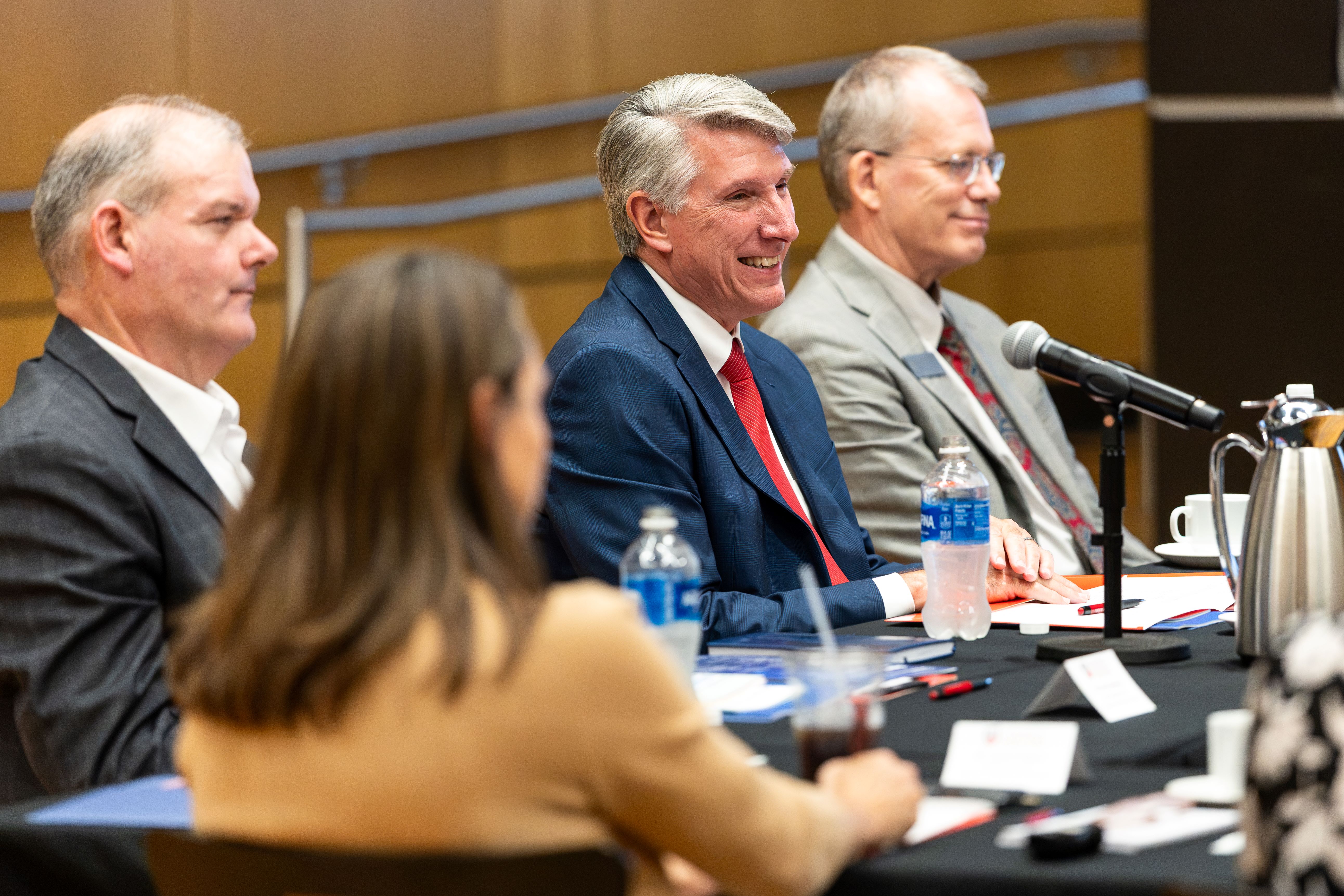 Three UCM officials are sitting at a table and smiling while listening to the UOS delegation. Everyone is in business attire and sitting at a table with a black tablecloth. They have various items in front of them, such as pens, notebooks, water bottles, and teacups. Another person sitting near the trio is seen from behind and is not in focus.