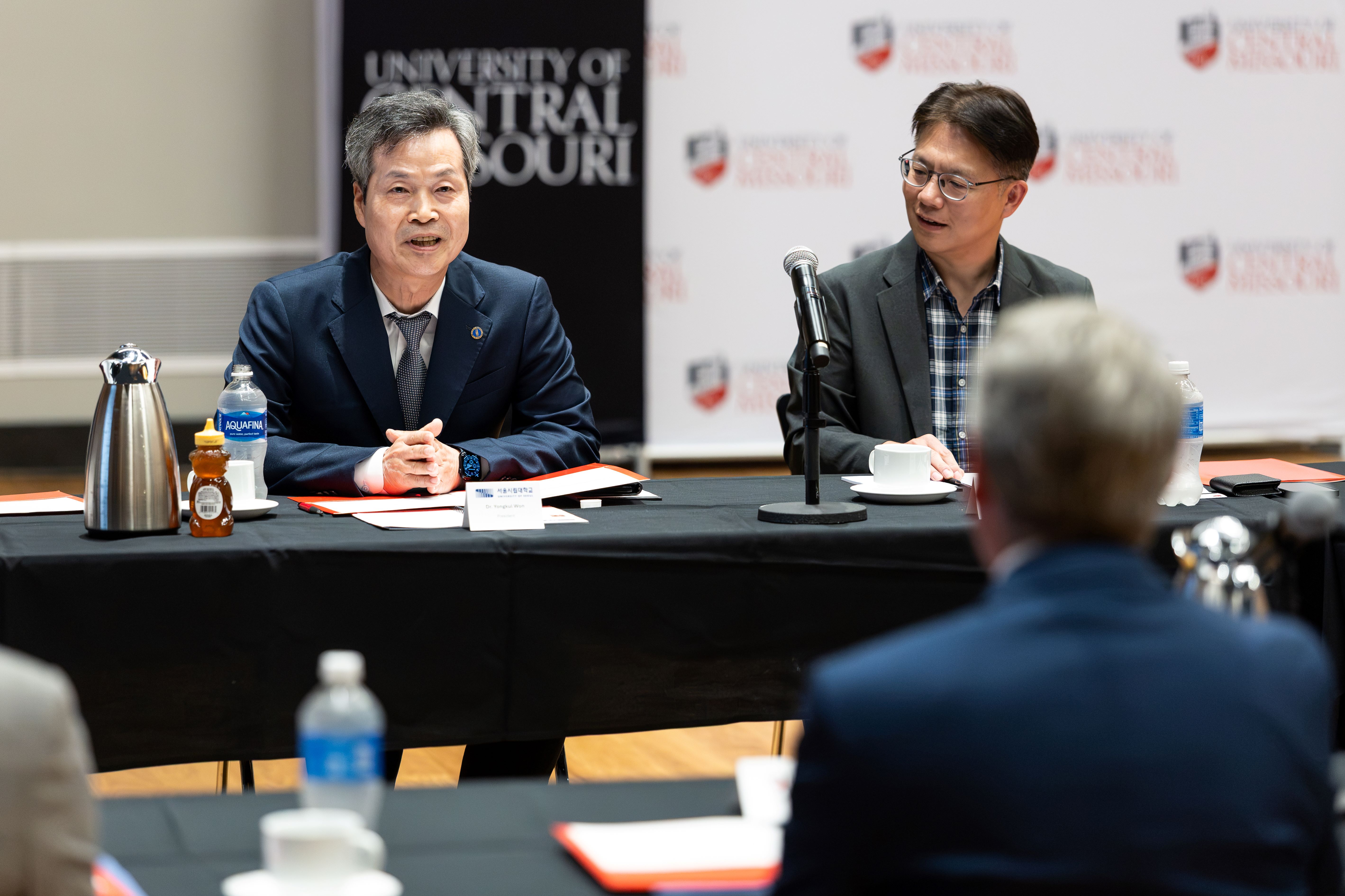 Two UOS leaders are sitting at a table with a black tablecloth and wearing suits. The UCM logo photo backdrop is seen in the distance behind them. One of them is talking while the other listens. Items are on the table such as pends, notebooks, folders, water bottles, a microphone on a stand, teacups, a silver metal tea kettle and a bottle of honey. A person from the UCM delegation sitting across the table is seen from behind and out of focus.