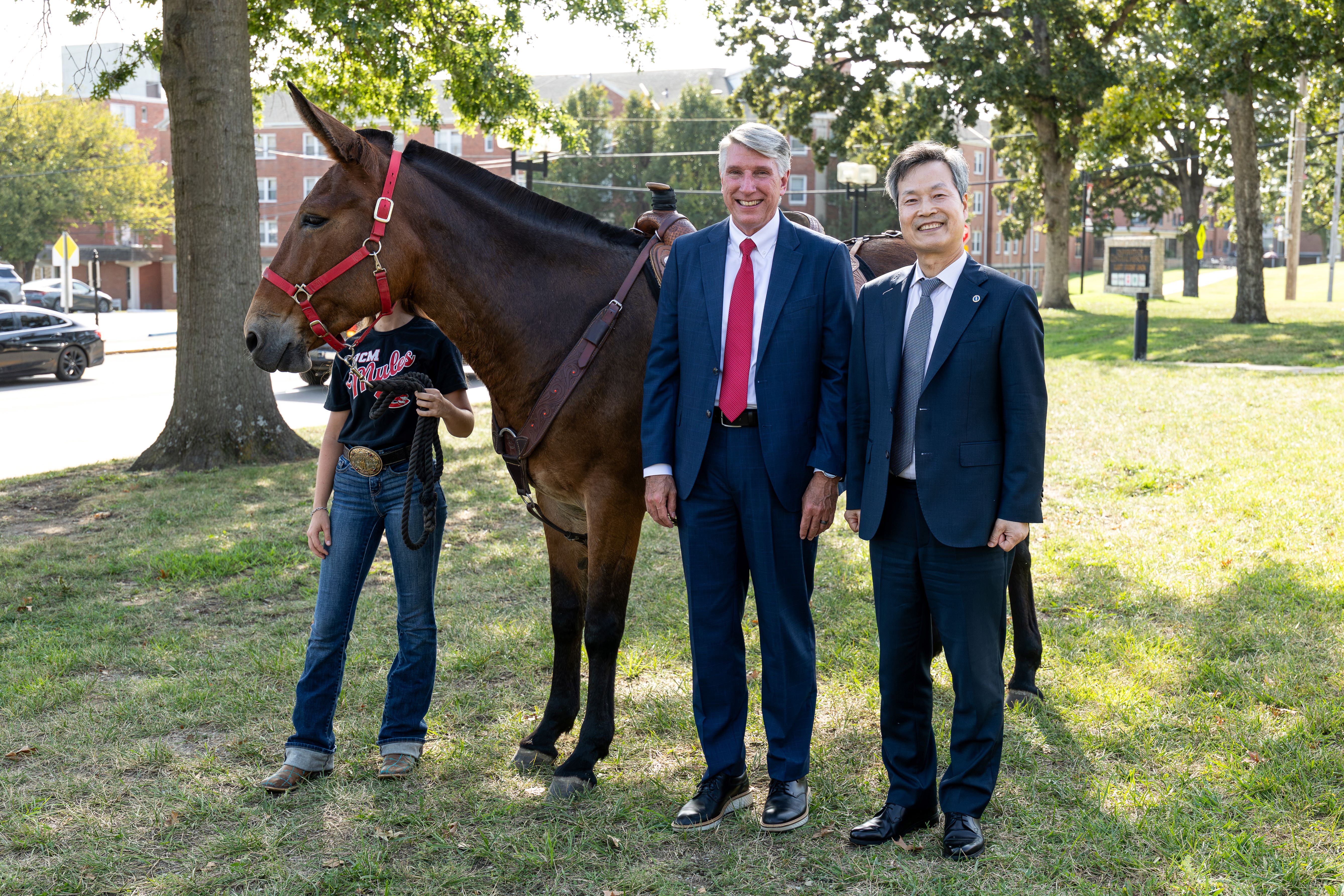 UCM President Best and UOS President Won are both wearing suits and ties. They are standing next to each other outside in a grassy area on the UCM campus. They are also standing next to Molly, UCM's live mule mascot, and a student member of the Mule Team who is wearing jeans and a black UCM T-shirt.