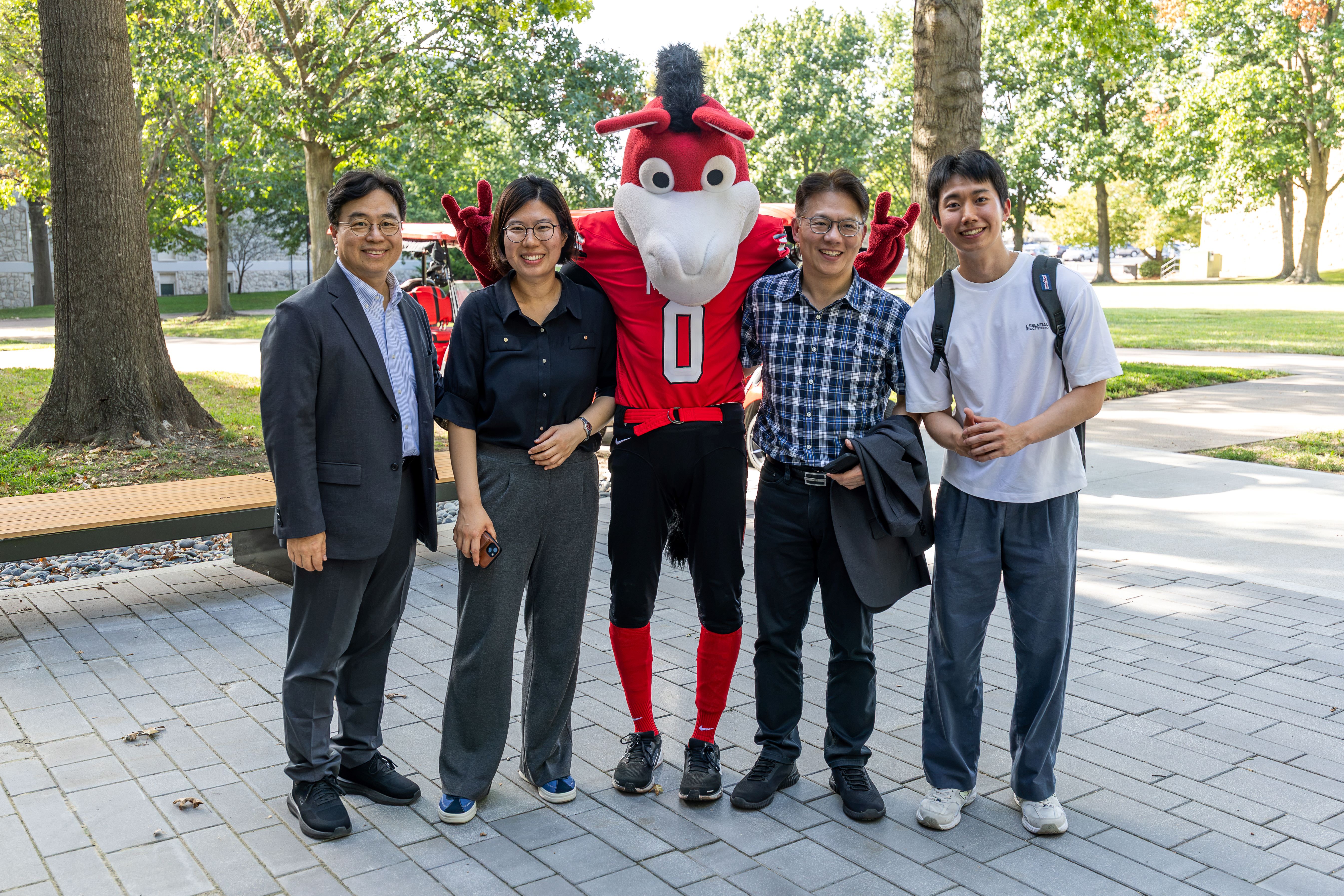 Three members of the UOS delegation and a UOS student studying at UCM pose for a photo with Mo the Mule, UCM's mascot. Two people stand on either side, with Mo in the middle. The group is outside on a sidewalk on the UCM campus with trees and a bench behind them. Mo is dressed in his Mules Football uniform and giving the Mule hand signal.