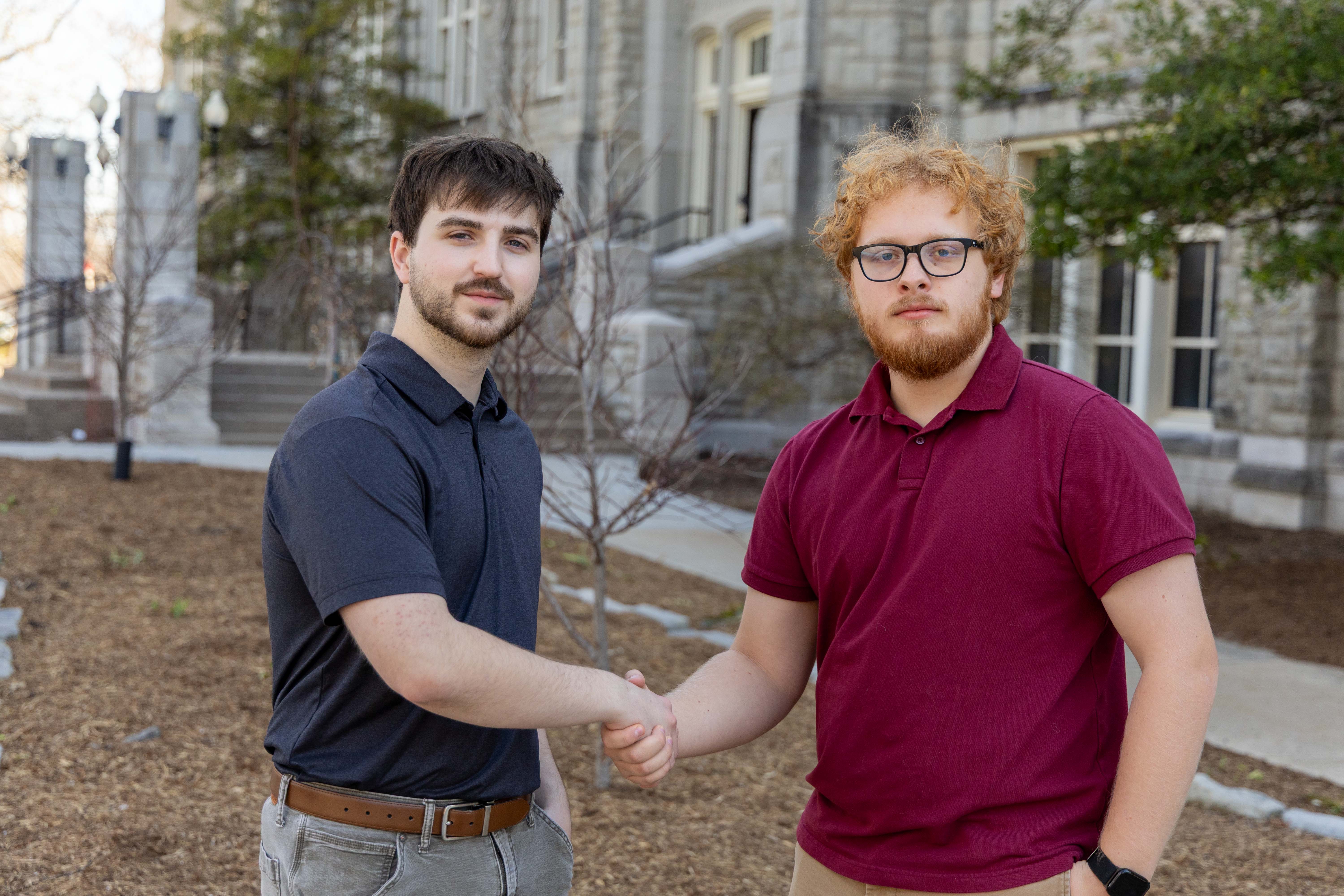 Two student workers shake hands while standing on a sidewalk with a stone building behind them.